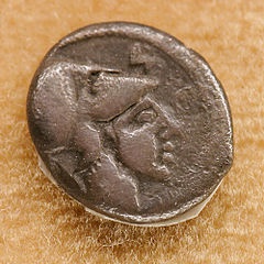 A close-up photograph of an ancient, weathered silver coin resting on a textured brown surface. The coin features a detailed…