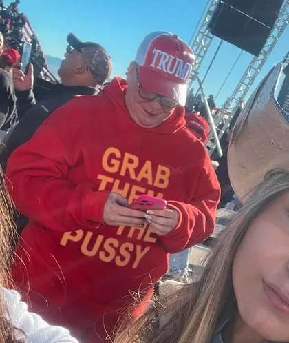 A man wearing a red 'TRUMP' cap and a red hoodie with the text 'GRAB THEM THE PUSSY' printed in yellow is looking down at…