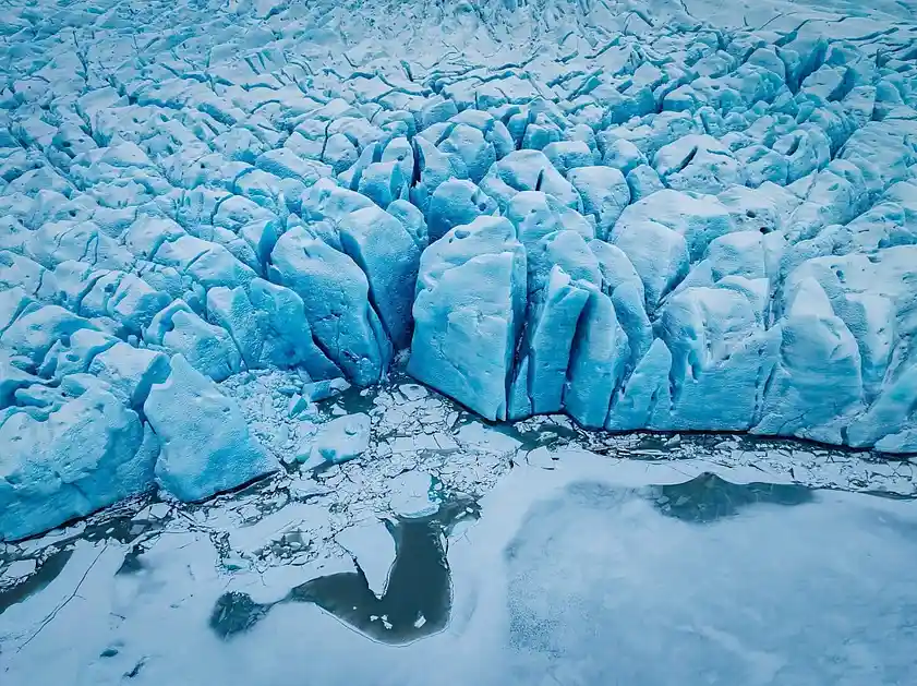 Aerial view of a glacier with jagged, blue-tinged ice formations meeting a cracked, icy surface with dark water pools. The…