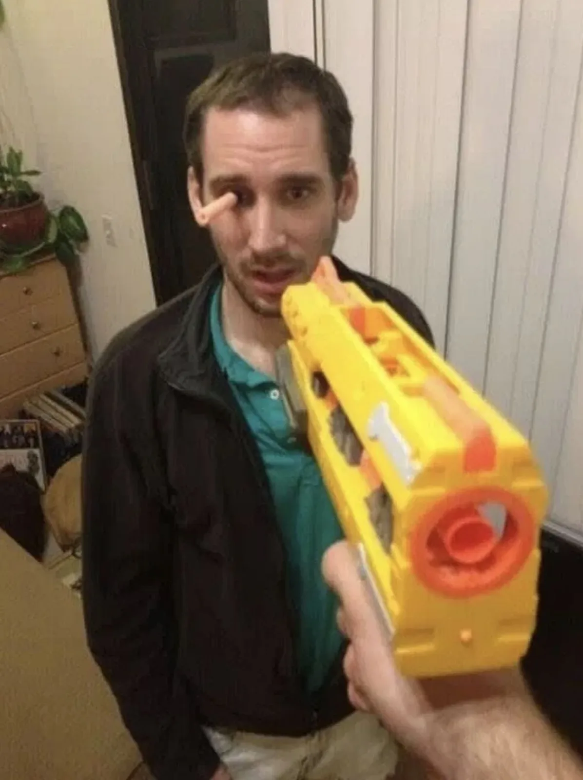 A man in a teal polo and black jacket stands indoors, looking directly at the camera with a surprised expression. A yellow…
