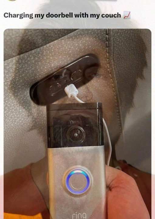A person holds a Ring doorbell camera, which is plugged into a charging port built into the armrest of a couch. The…