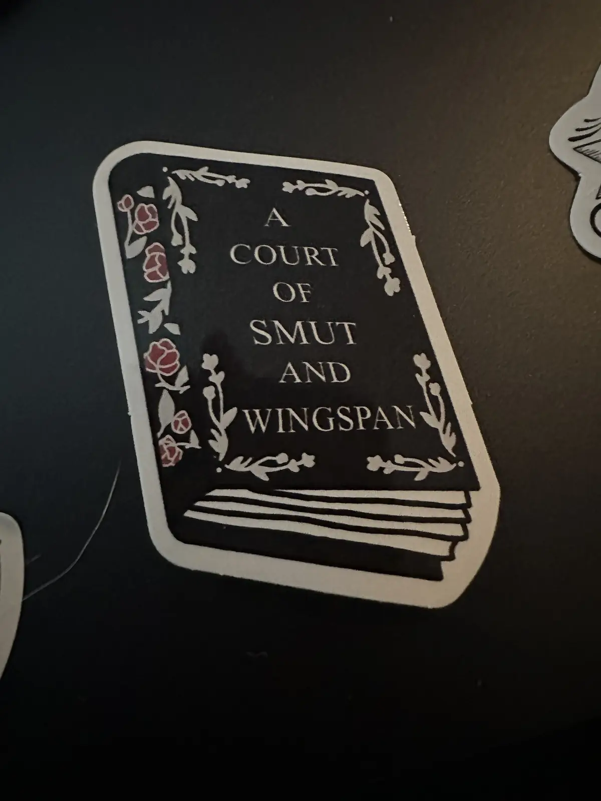 A black sticker depicts a book with ornate white floral borders and red roses, its title altered to 'A COURT OF SMUT AND…