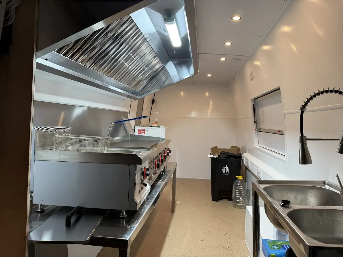 A clean, stainless steel commercial kitchen interior with a large fryer and griddle under a vent hood, a double sink with a…