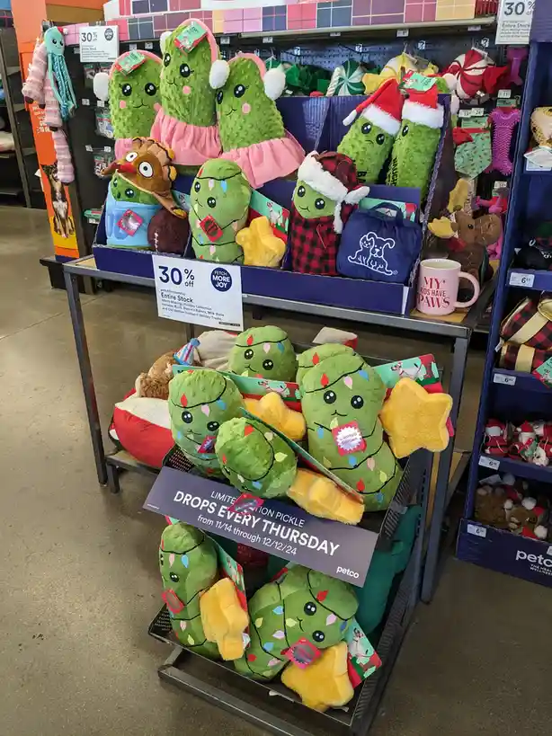 A Petco store display features plush holiday-themed pickle toys dressed in festive outfits like Santa hats and Christmas…