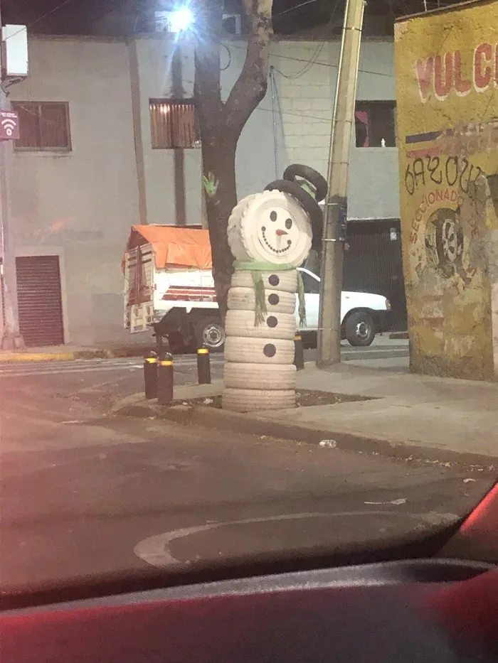 A snowman made of stacked tires stands on a sidewalk at night, wearing a tire hat and scarf, with a painted smiling face…