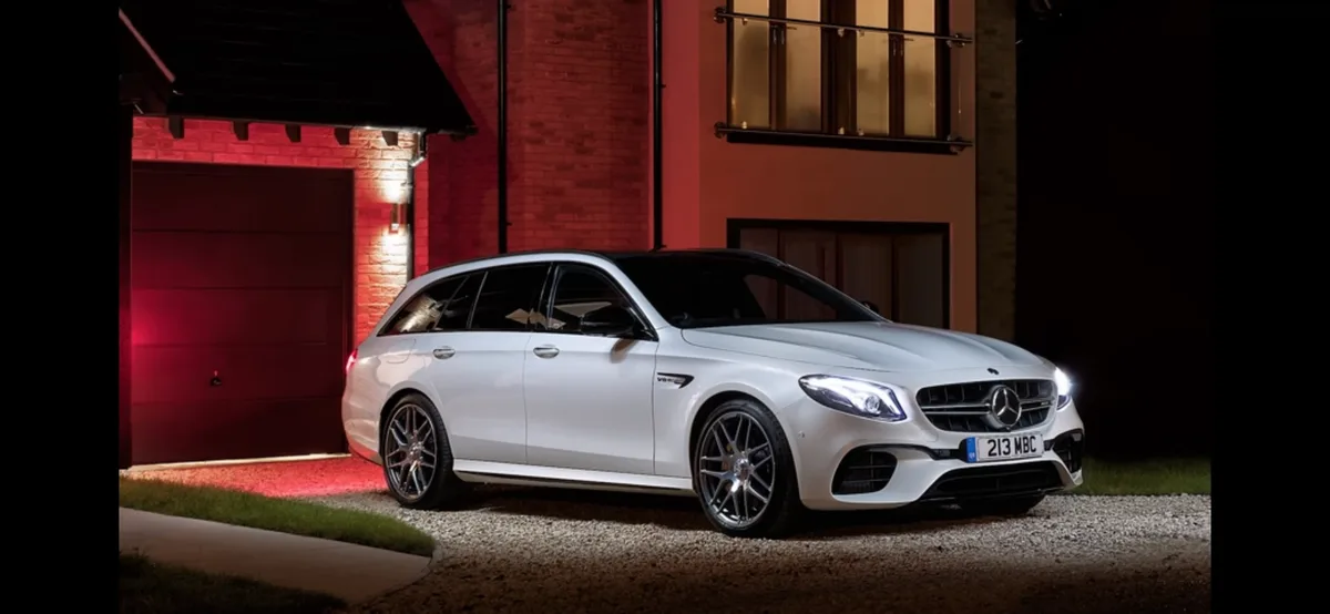 A white Mercedes-AMG E63 S Estate is parked on a gravel driveway at night in front of a modern brick house with red…