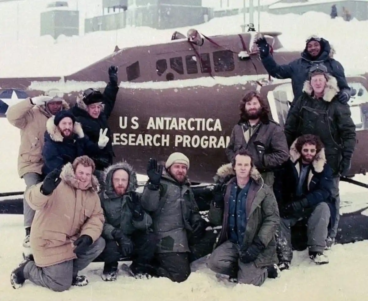 A vintage photo shows a group of men in heavy winter gear posing cheerfully in front of a brown airplane covered in snow…