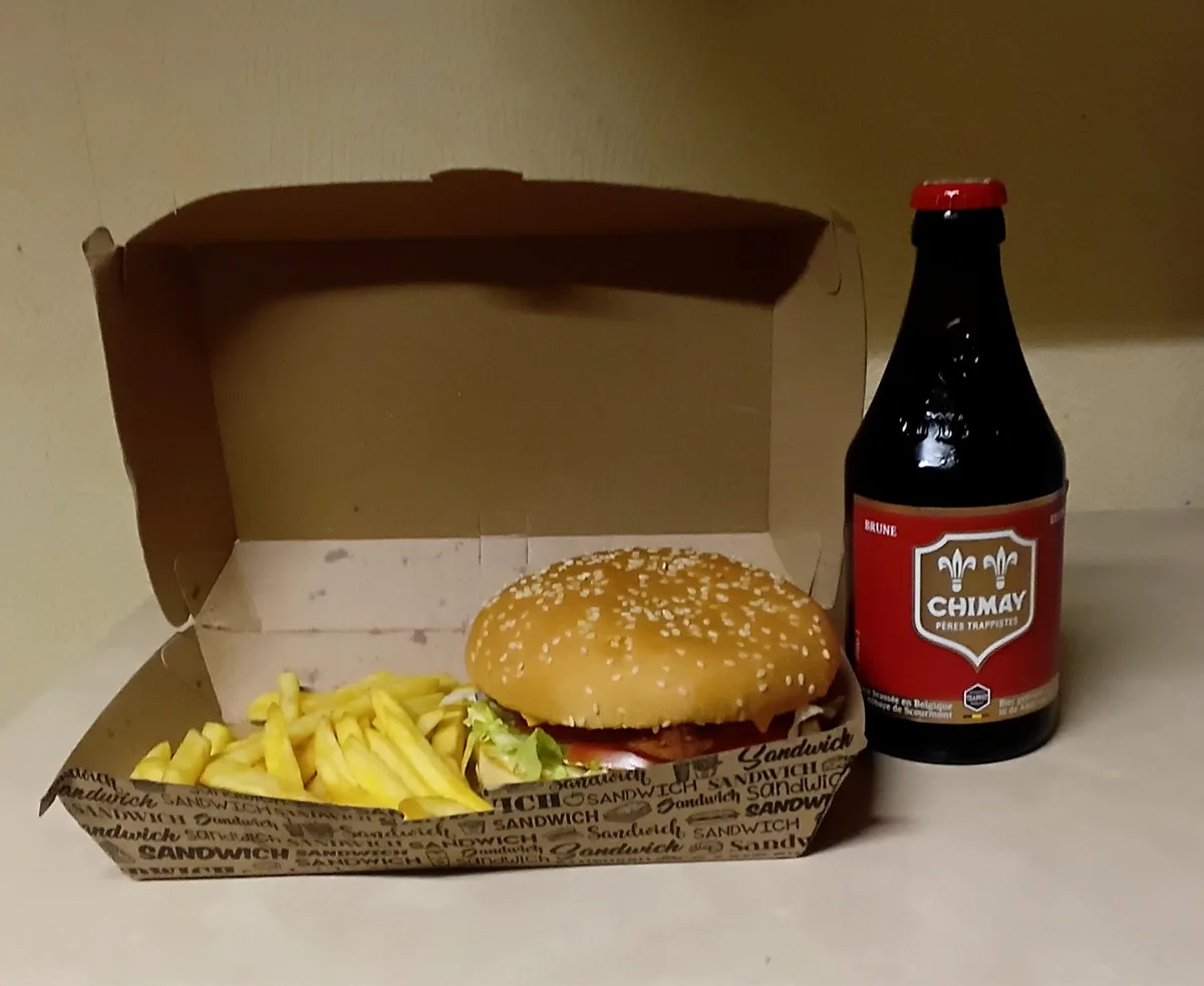 A burger with fries sits in a cardboard box printed with the word 'sandwich' repeatedly, next to a bottle of Chimay beer…