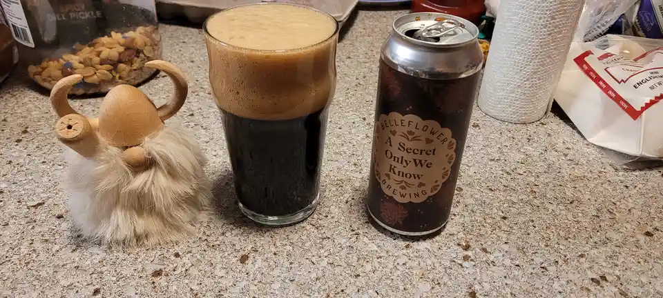 A wooden Viking-style figurine with horns and a fluffy beard sits next to a glass of dark beer with a thick tan head and an…