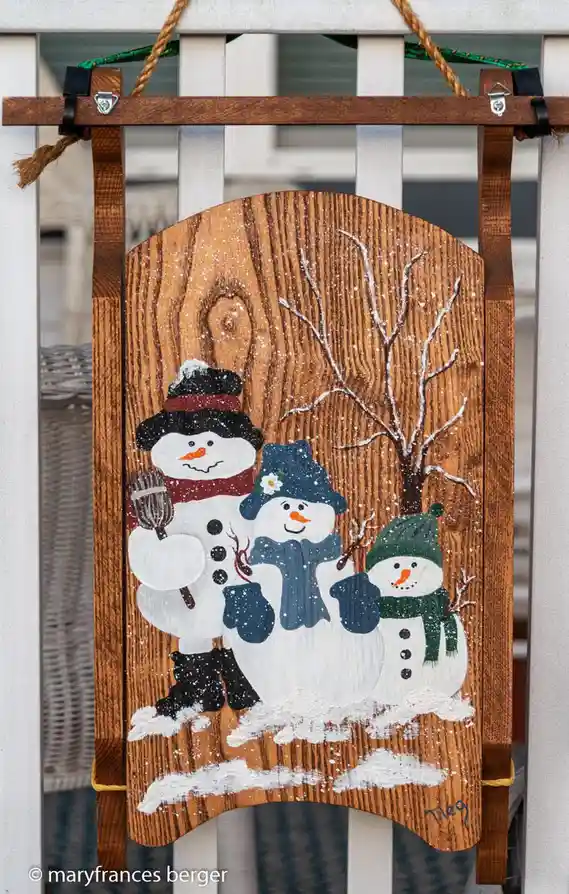 A wooden decorative sign painted with three snowmen standing in a snowy scene with a bare tree, hanging on a white railing…