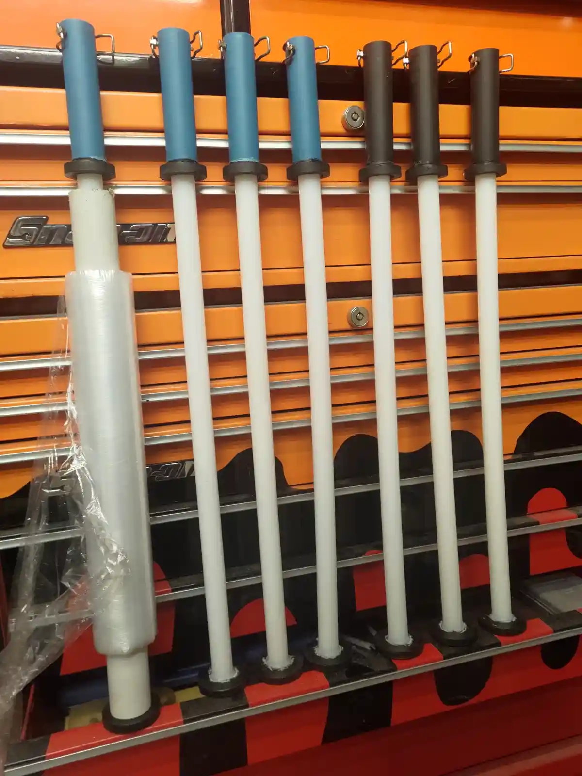 Seven long white tools with blue or black handles hang neatly on an orange and black tool cabinet. The leftmost tool is…