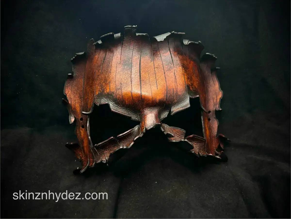 A dark, textured leather mask with jagged, irregular edges and eye holes, resembling a skull or demonic face, is displayed…