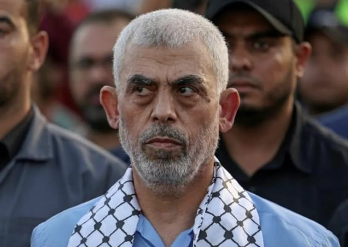 A man with gray hair and a beard, wearing a blue shirt and a black-and-white checkered keffiyeh, looks sideways with a…