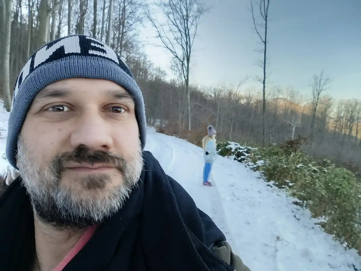 A man with a gray beard and a blue and white knit beanie takes a selfie on a snowy path in a forest, looking directly at the…