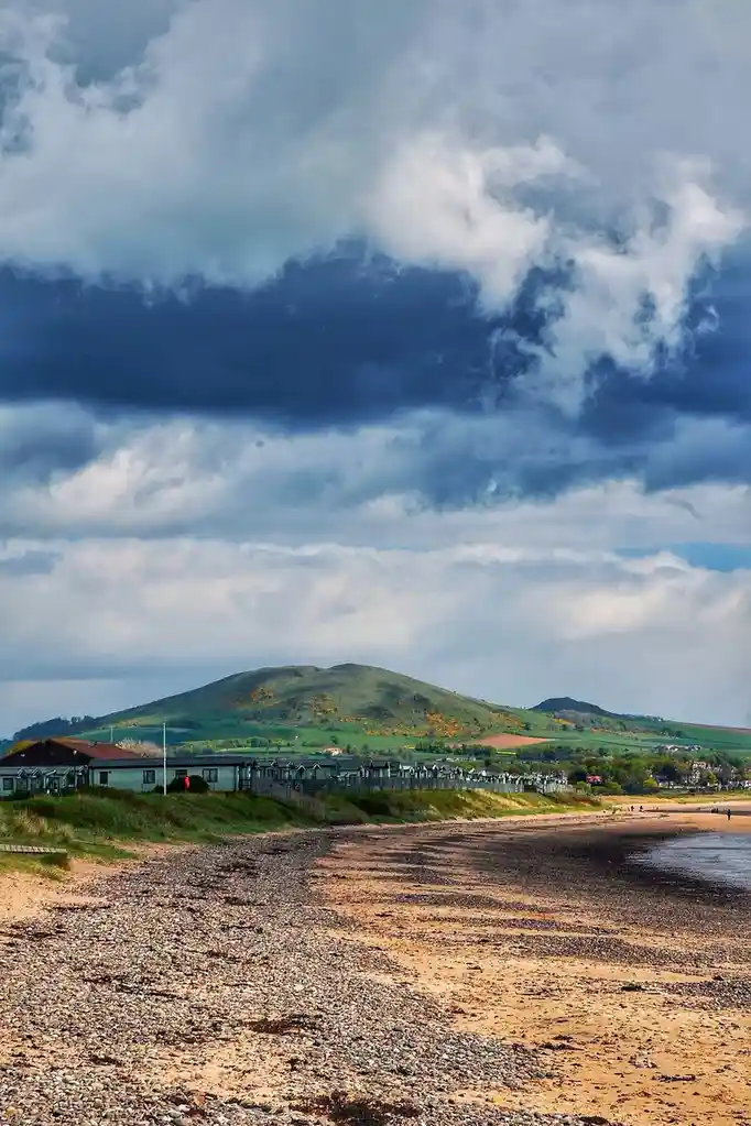 A scenic beach photo with a pebbly shore, green hills in the background, and dramatic clouds overhead. Overlaid text reads…