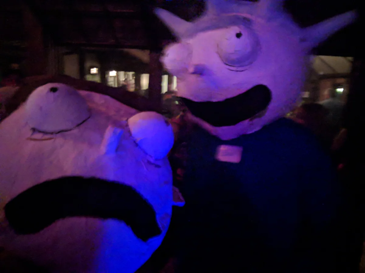 Two large, cartoonish monster costumes with wide, open mouths and simple eyes are shown in a dimly lit, purple-hued…