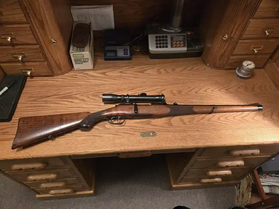 A wooden rifle with a scope rests horizontally on a wooden desk with drawers, surrounded by reloading equipment including a…