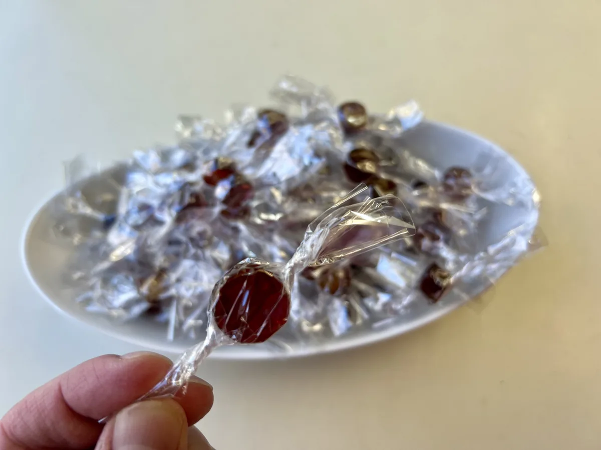 A hand holds a single, individually wrapped red lollipop in the foreground, with a white plate piled high with dozens of…