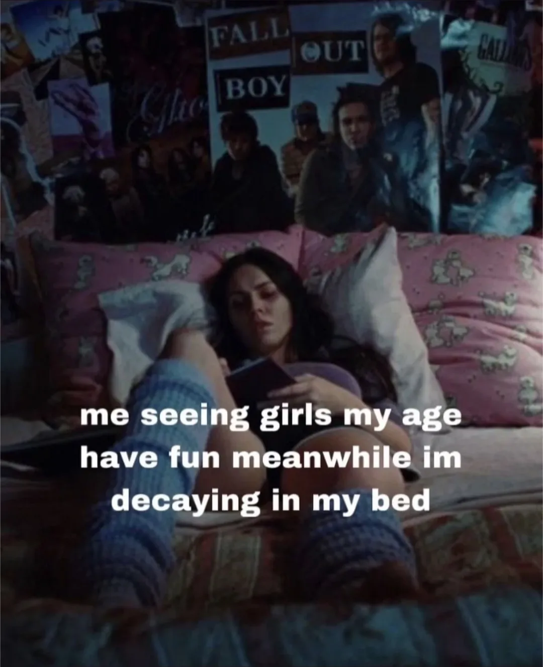 A young woman with dark hair lies in bed surrounded by posters, including one for Fall Out Boy, looking bored while holding…