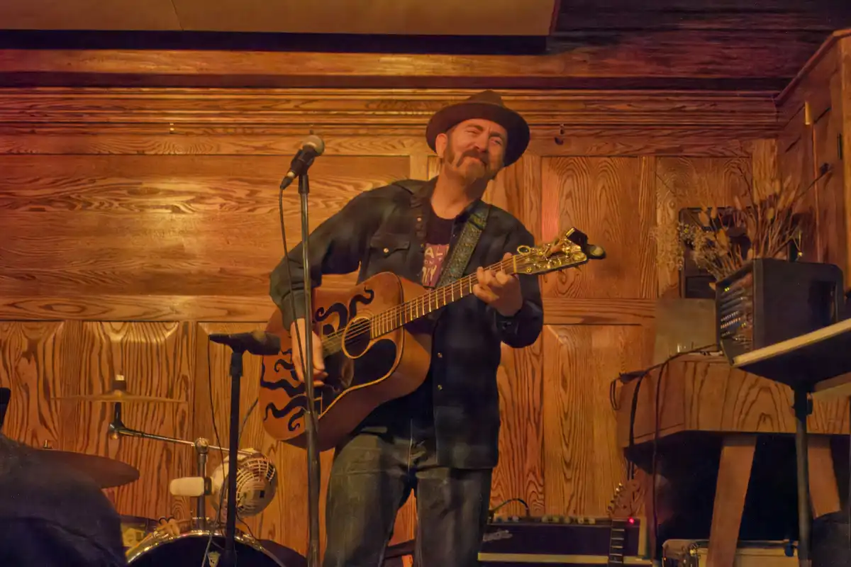 A man with a mustache and hat plays an acoustic guitar on a wooden-paneled stage with a microphone and drum kit visible. The…