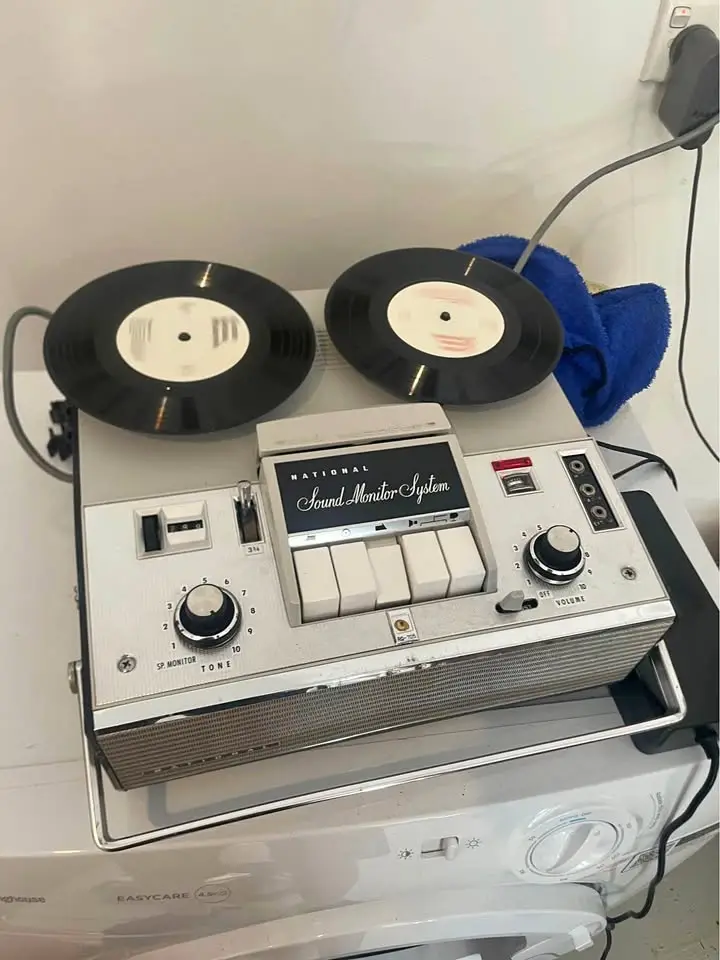 A vintage silver 'National Sound Monitor System' reel-to-reel tape recorder sits atop a white washing machine, with two…