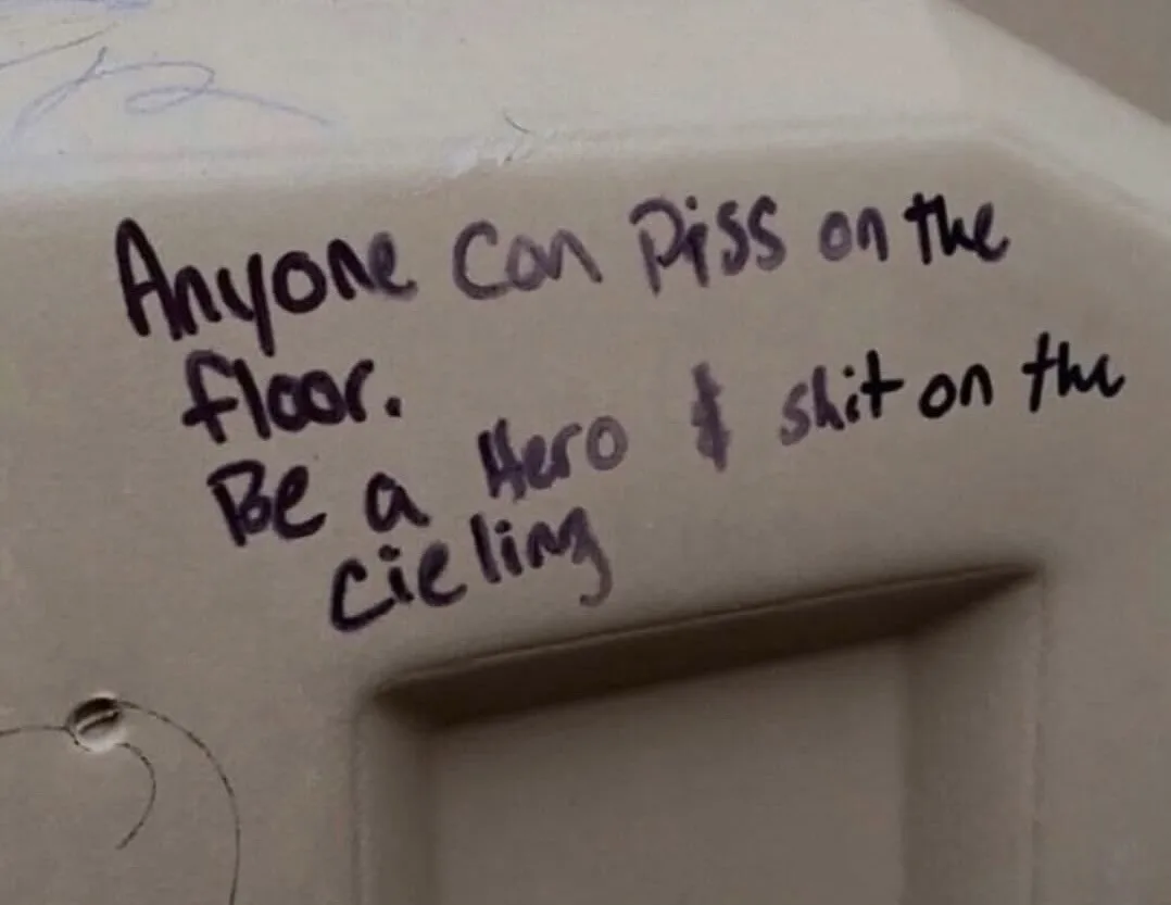 A close-up of handwritten text in dark ink on a white bathroom wall near a doorway. The text reads: 'Anyone can piss on the…