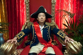 A man in a pirate costume with a tricorn hat sits on a golden throne, arms outstretched, against a backdrop of red curtains…