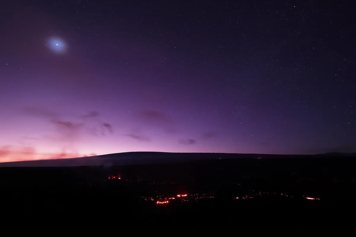A starry night sky with a bright, glowing moon-like object in the upper left, above a dark landscape dotted with distant red…