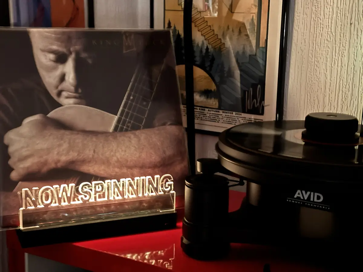 A close-up shot shows a vinyl record player with a record on its platter, next to a glowing 'NOW SPINNING' sign. Behind them…