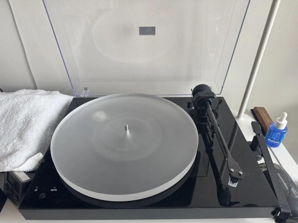 A black turntable with a clear dust cover is shown, its platter spinning empty with no vinyl record on it. The tonearm is…