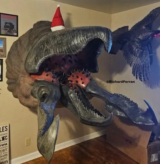 A large, realistic taxidermy-style dinosaur head mounted on a beige wall, wearing a small red and white Santa hat. To its…