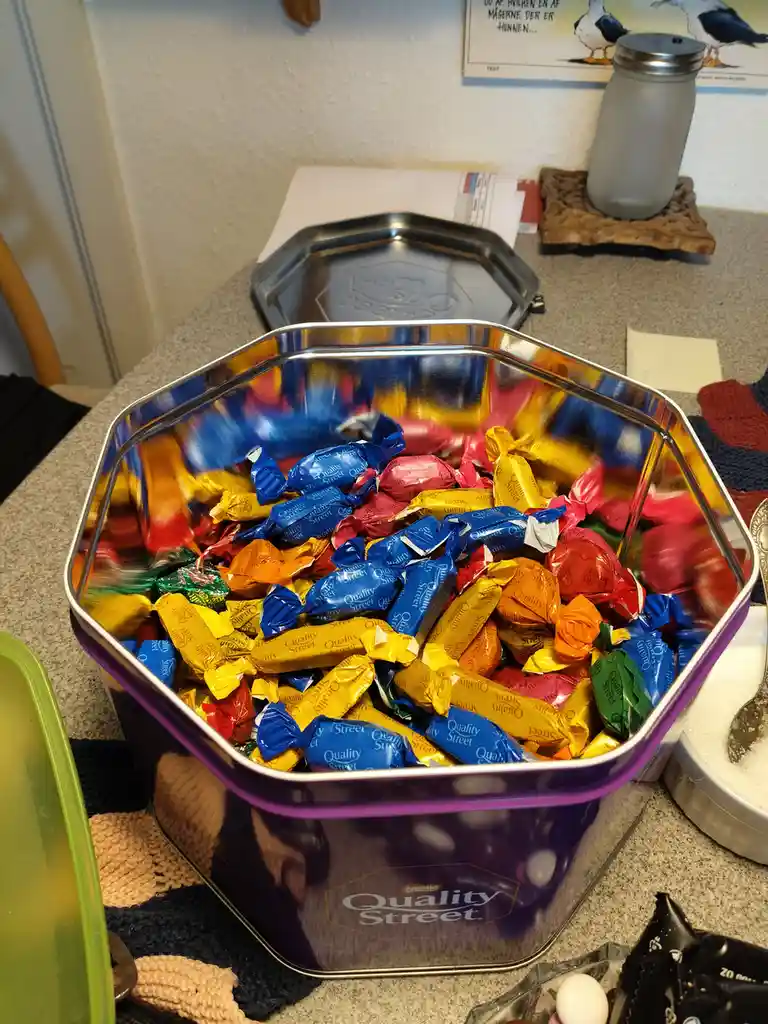 A purple hexagonal tin labeled 'Quality Street' is filled with colorful individually wrapped candies in blue, yellow, red…