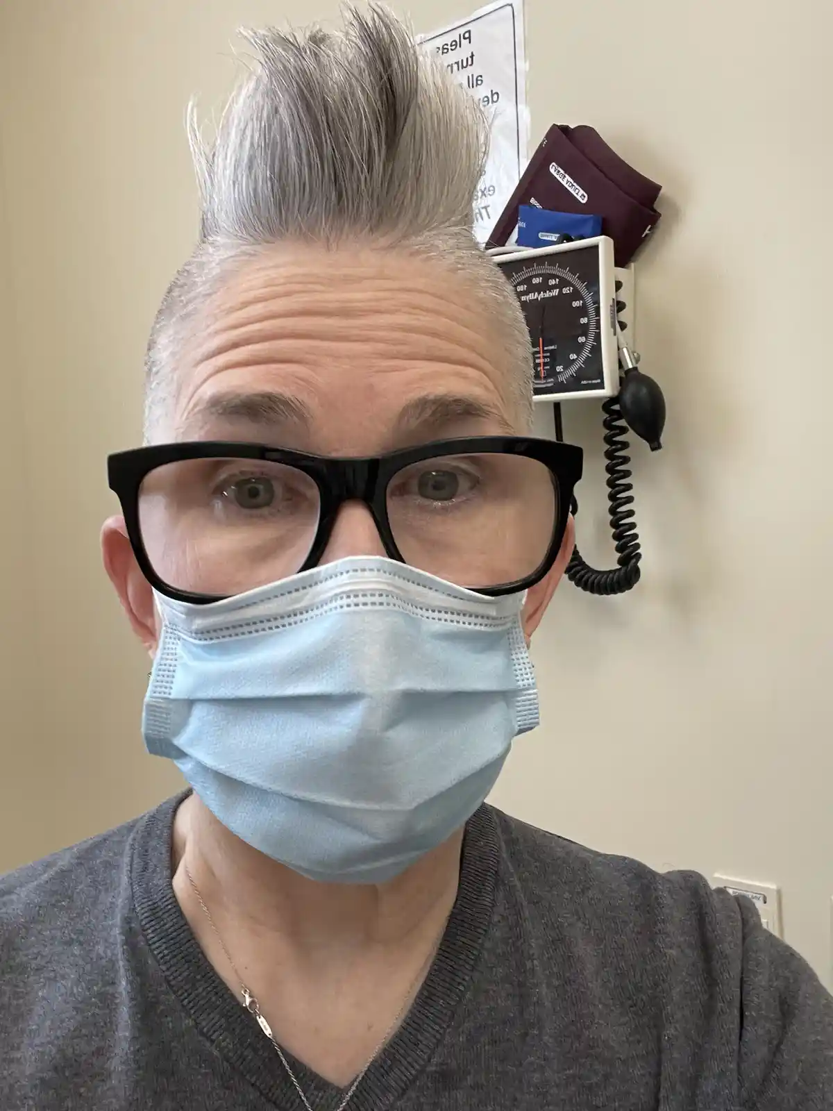 A person with short, spiky gray hair and large black-rimmed glasses wears a light blue surgical mask while looking directly…