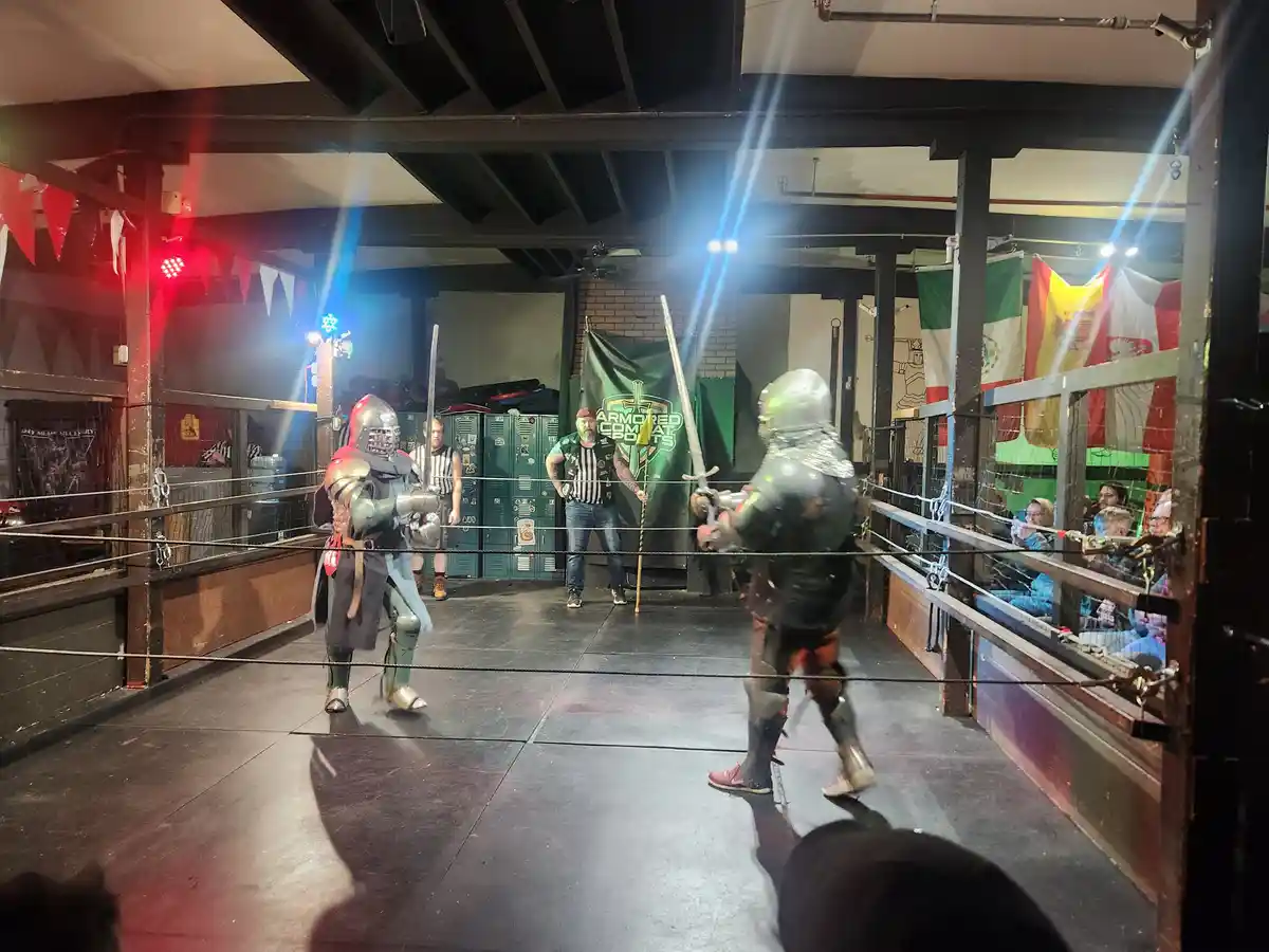 Two people in full medieval-style armor are facing each other in a fenced arena, holding swords, with a referee and…