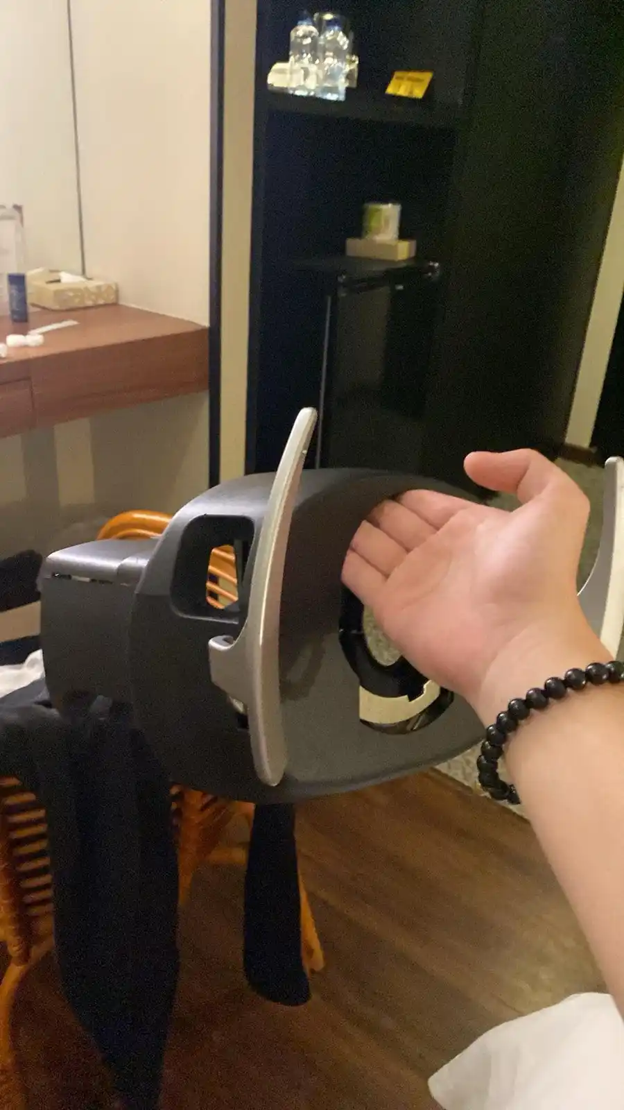 A person's hand is shown resting on the back of a chair in a hotel room, with a mini fridge and desk visible in the…