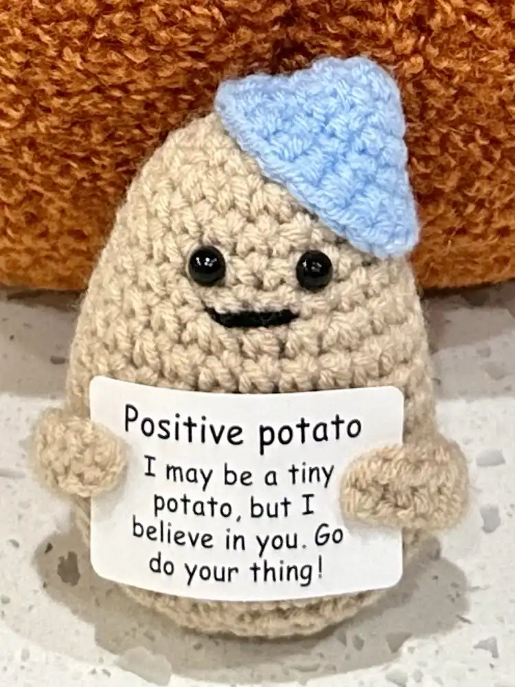 A handmade crochet potato plushie wearing a small blue crocheted hat and holding a white sign. The sign reads 'Positive…