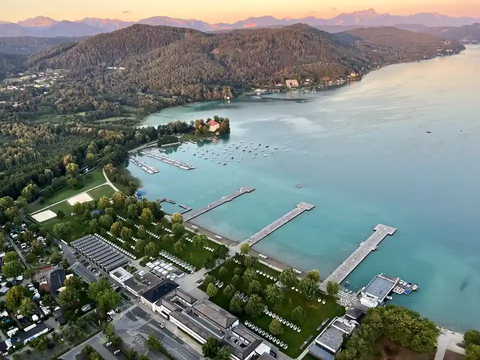 An aerial photo of a serene lake surrounded by forested hills and mountains at sunset, featuring docks, boats, and a resort…