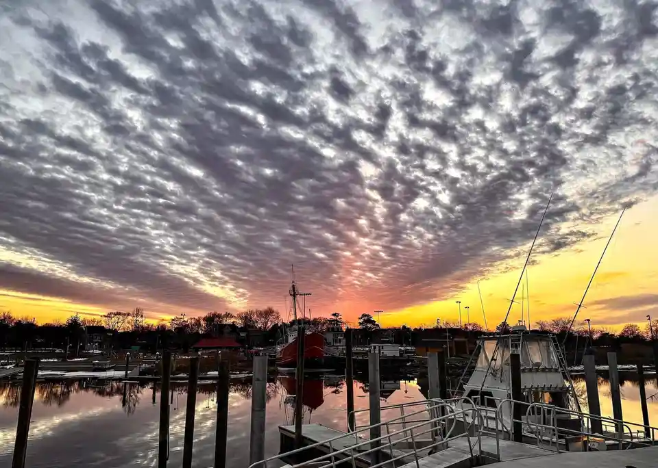 A serene harbor at sunset with boats docked, featuring a dramatic sky filled with textured clouds reflecting in calm water…