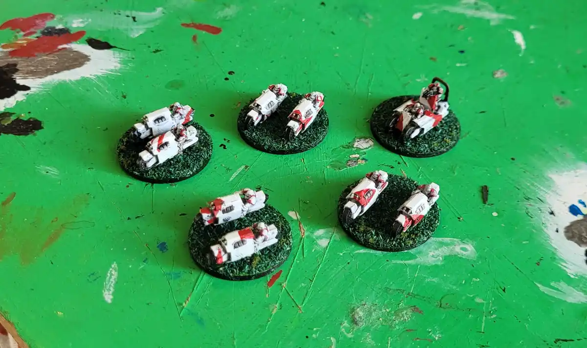 Several painted miniature motorcycle riders on circular green bases are arranged on a bright green, paint-splattered…