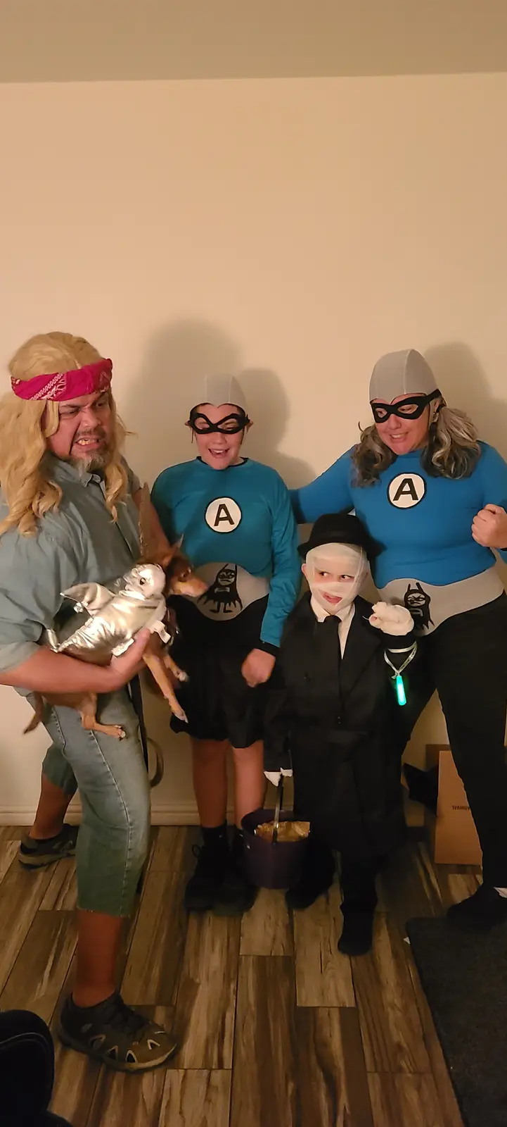 Four people and a dog pose indoors in costumes: one man as a blonde-haired character with a pink bandana holding a dog…