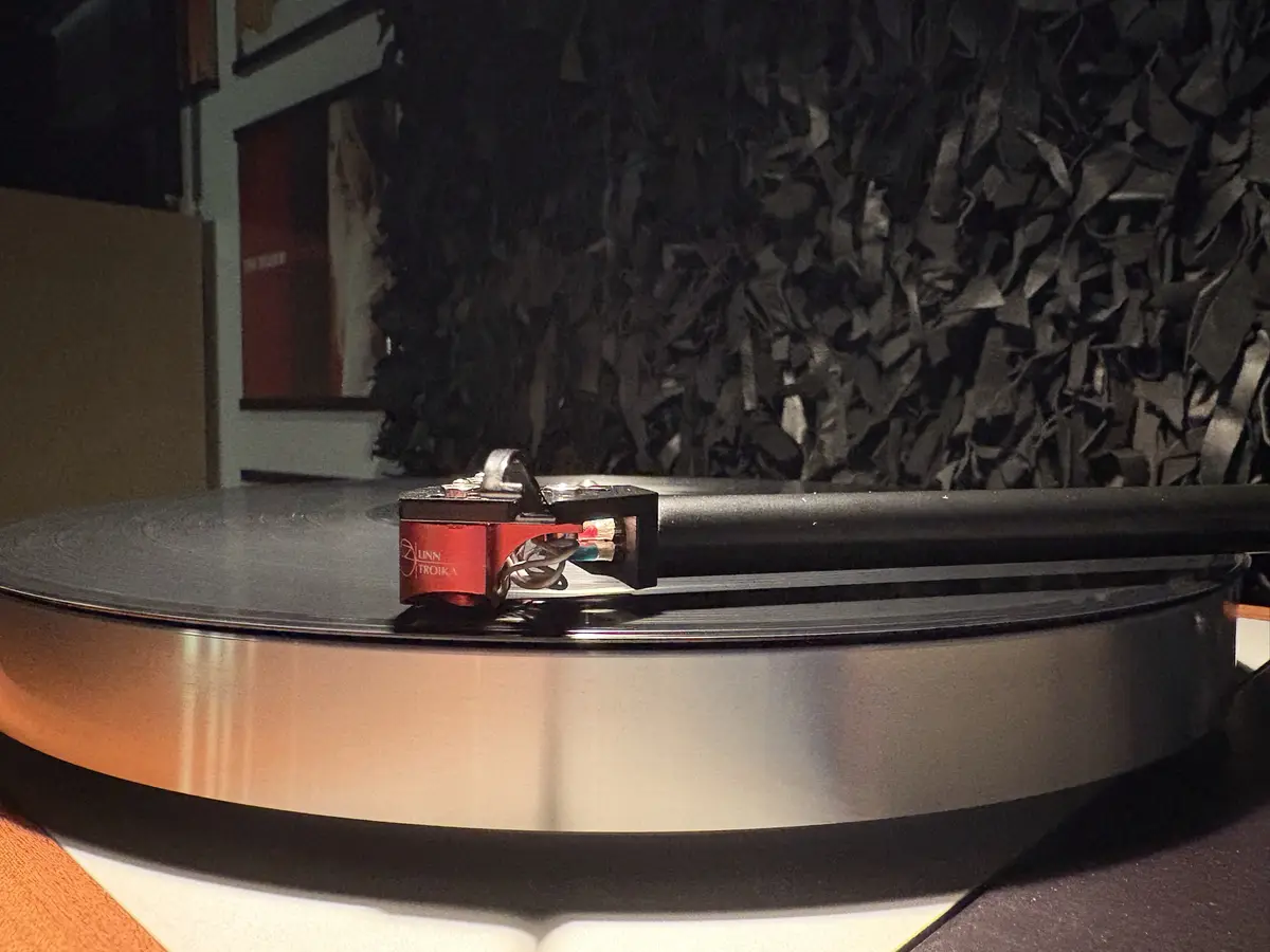 A close-up photo of a turntable with a red Linn Troika cartridge resting on a spinning vinyl record. The background features…