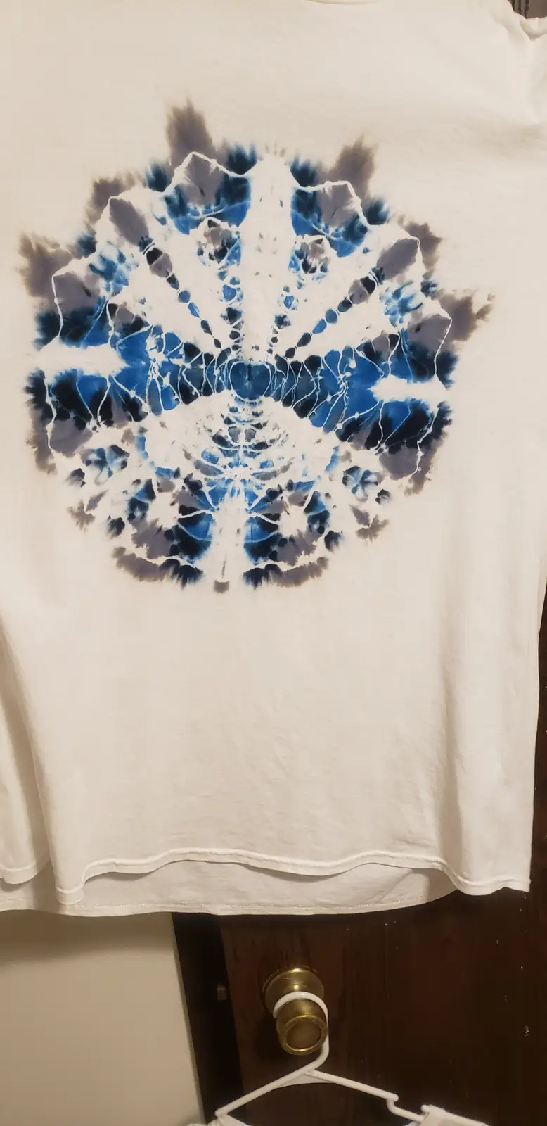 A white t-shirt with a circular blue and white tie-dye pattern resembling a spider web hangs on a white hanger against a…