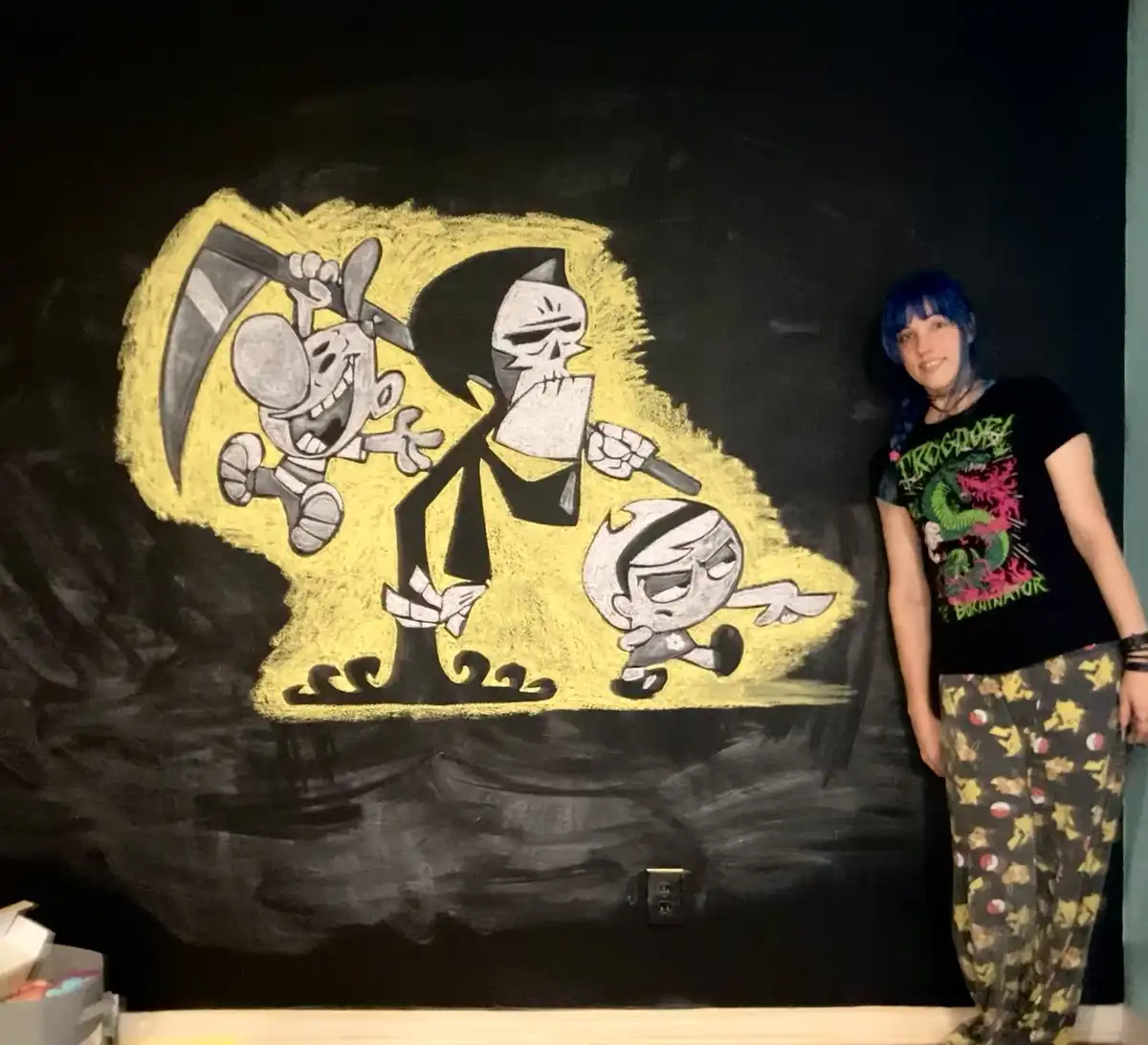 A person with blue hair stands beside a large chalk drawing on a black wall, depicting characters from 'The Grim Adventures…