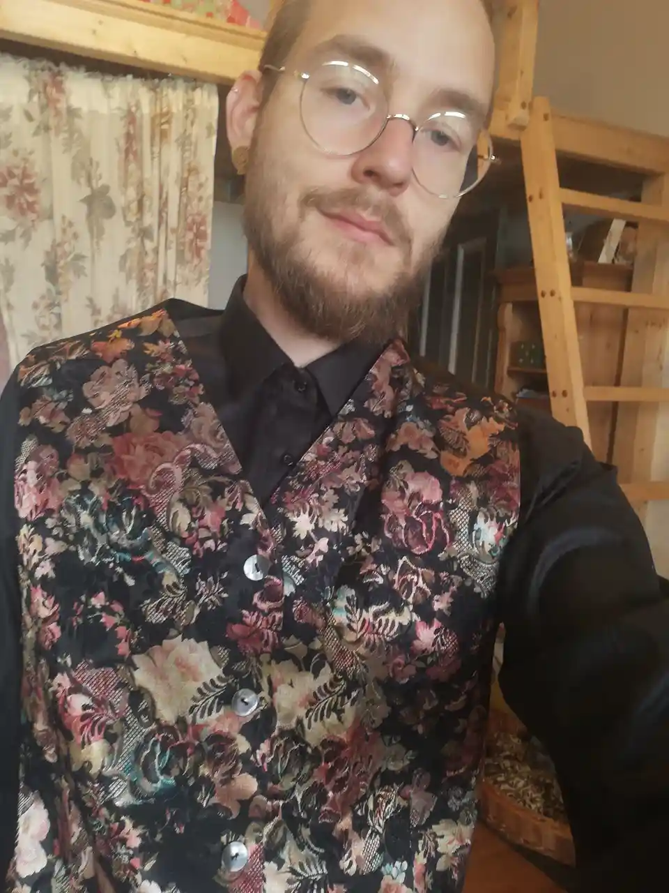 A man with a beard and round glasses takes a selfie wearing a black shirt under a very ornate, colorful floral-patterned…