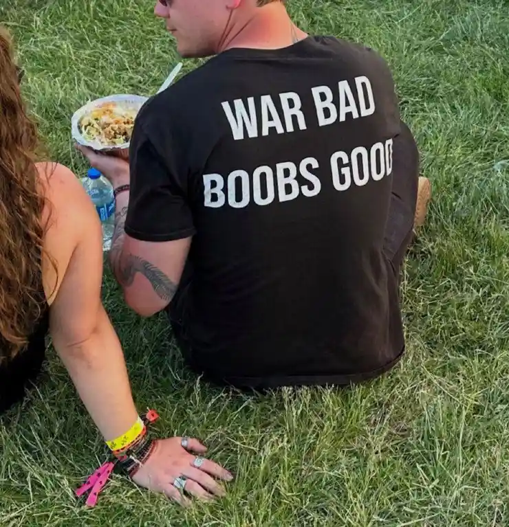 A man is seen from behind, sitting on grass with a woman beside him, wearing a black t-shirt with the text 'WAR BAD BOOBS…