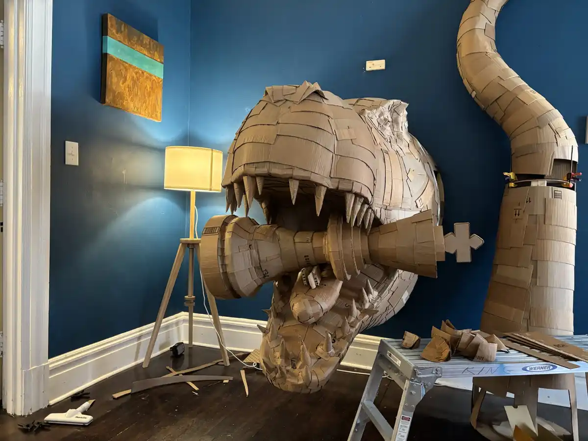 A large, intricate cardboard sculpture of a dragon's head with an open mouth and sharp teeth is positioned in a room with…