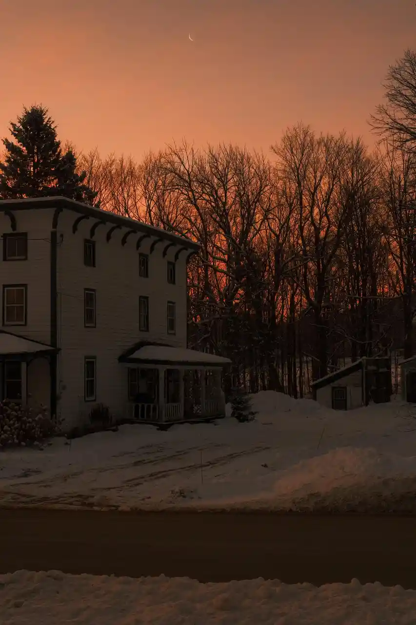 A snowy winter scene at dusk or dawn, featuring a white house with a porch, snow-covered ground, bare trees, and a crescent…