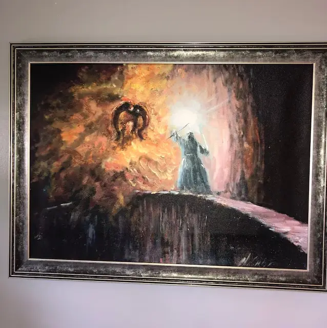 A framed painting depicts a dark, dramatic scene with a robed figure holding a glowing sword, facing a fiery, horned demonic…