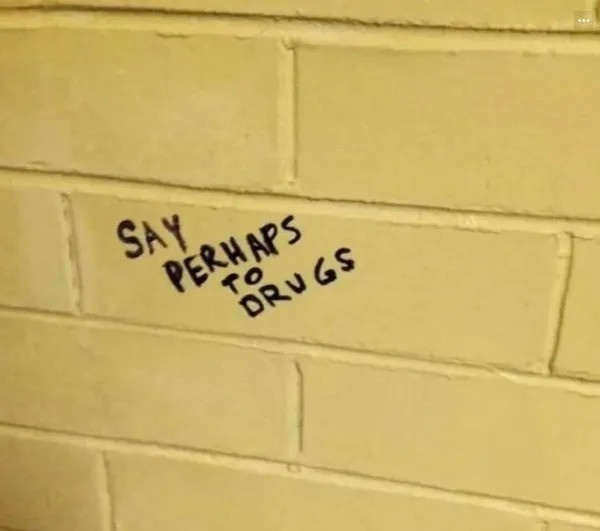 A close-up of a pale yellow brick wall with handwritten text in black marker that reads 'SAY PERHAPS TO DRUGS', written…