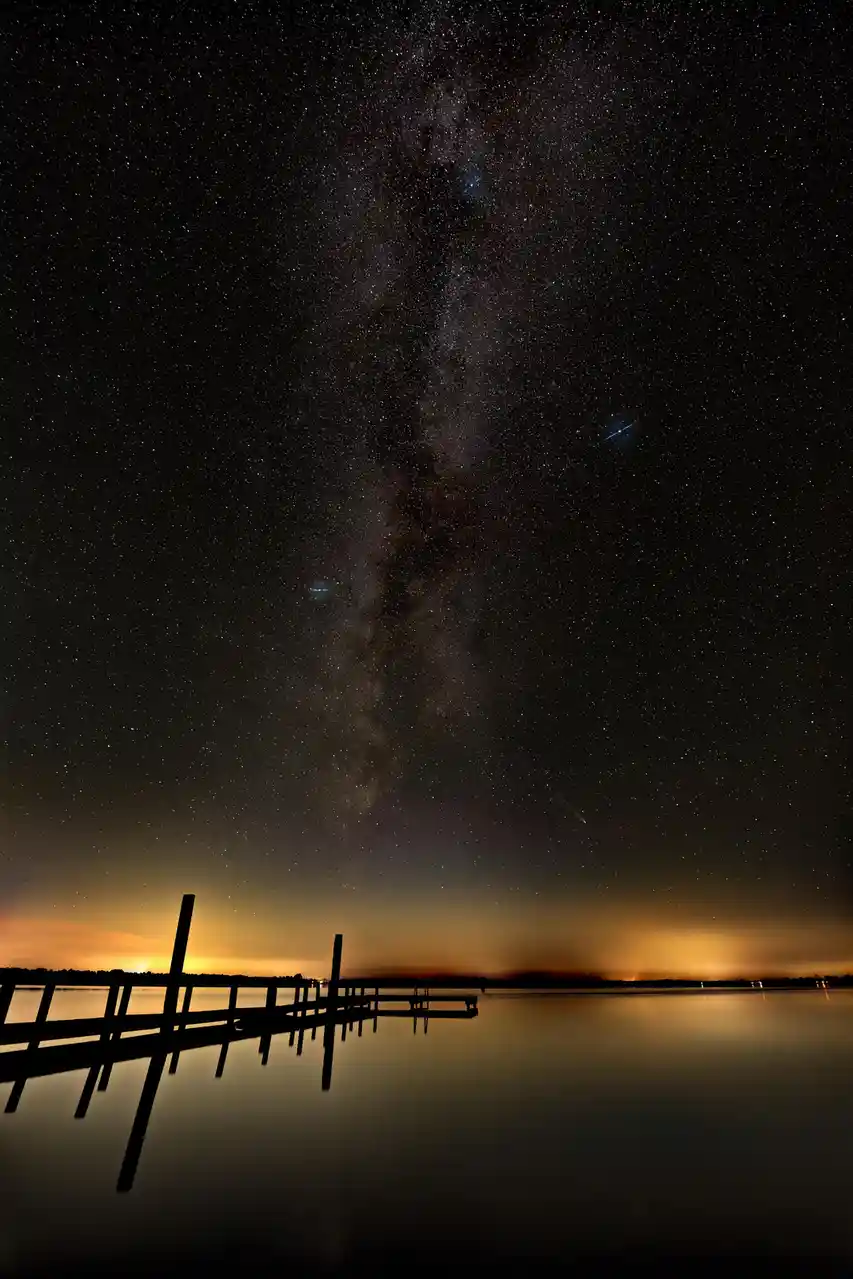A serene night sky photograph shows the Milky Way galaxy stretching vertically across a dark, star-filled sky, with a warm…